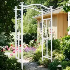 White metal arbour with climbing roses on a garden path beside a cabin